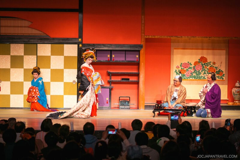 A traditional Japanese stage performance at Edo Wonderland Nikko Edomura shows an elegantly dressed woman in a kimono walking, accompanied by another woman, while two men in costumes sit on the floor. An audience watches attentively.