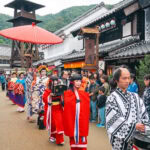 Edo Wonderland Nikko Edomura (江戸ワンダーランド日光江戸村) – Step Into Japan’s Edo Period A traditional Japanese parade at Edo Wonderland Nikko Edomura features people in colorful kimonos and costumes walking down a street lined with old-style buildings, as onlookers watch and take pictures. A large red umbrella is held above one participant.