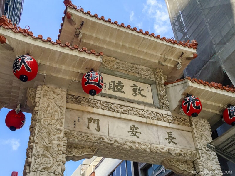 A traditional Chinese-style entrance gate with intricate carvings and red lanterns, set between modern buildings under a blue sky in Kobe Chinatown. Chinese characters are engraved on the gate.