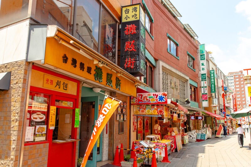 A lively street scene in Kobe Chinatown features colorful storefronts, restaurant signs with Japanese text, outdoor displays of food items, and a few people walking under sunny skies.