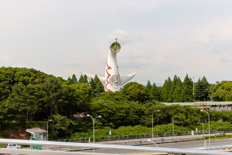 A large white sculpture with red patterns and three protruding arms, known as the Tower of the Sun, stands among green trees near Osaka Expocity under a partly cloudy sky in a park setting with roads in the foreground.