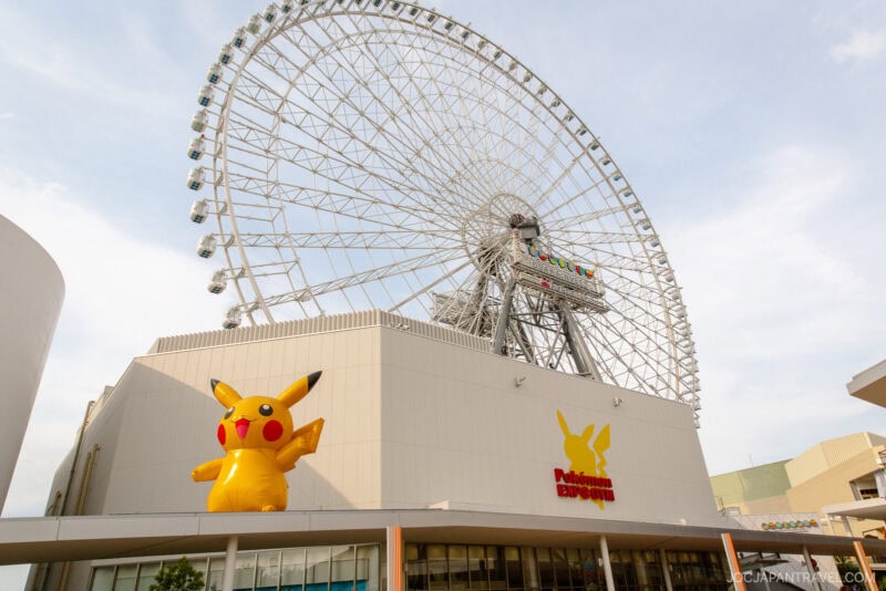 A large Ferris wheel rises behind the Pokémon Expo Gym building at Osaka Expocity, featuring a giant inflatable Pikachu and the yellow Pokémon logo on its facade. The sky is mostly clear with a few clouds.