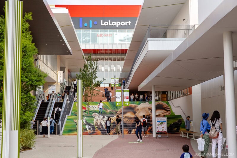 Modern shopping mall entrance labeled Lalaport at Osaka Expocity with people on escalators and walking; colorful mural of animals decorates the stairs; trees and outdoor seating visible on the right.