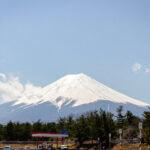 Exploring Fuji-Q Highland (富士急ハイランド) – Thrills at the Foot of Mt. Fuji Snow-capped Mount Fuji rises under a clear blue sky, with the Fuji-Q Highland roller coaster on the left, trees at the base, and a small gas station and parked cars in the foreground.