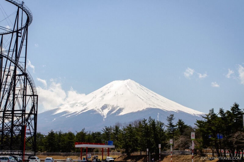 Snow-capped Mount Fuji rises under a clear blue sky, with the Fuji-Q Highland roller coaster on the left, trees at the base, and a small gas station and parked cars in the foreground.