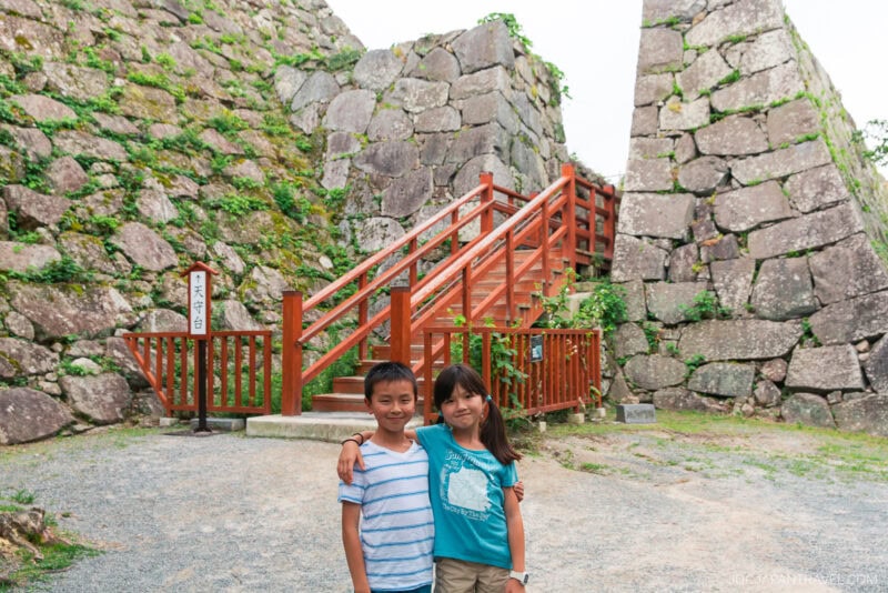 Two children stand smiling in front of a wooden staircase and fence, surrounded by large stone walls covered in patches of greenery at the historic Fukuoka Castle outdoor site.