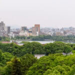Fukuoka Castle (福岡城) – Ruins, Views, and Seasonal Beauty A city skyline with mid-rise buildings and cranes rises behind a lush green park, lake, and the historic Fukuoka Castle, under a cloudy sky. Dense trees fill the foreground.