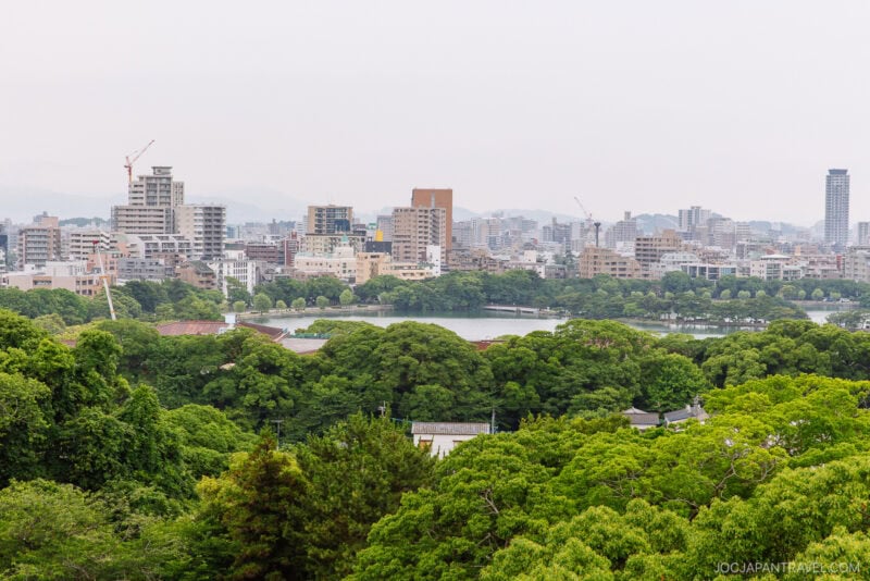 A city skyline with mid-rise buildings and cranes rises behind a lush green park, lake, and the historic Fukuoka Castle, under a cloudy sky. Dense trees fill the foreground.