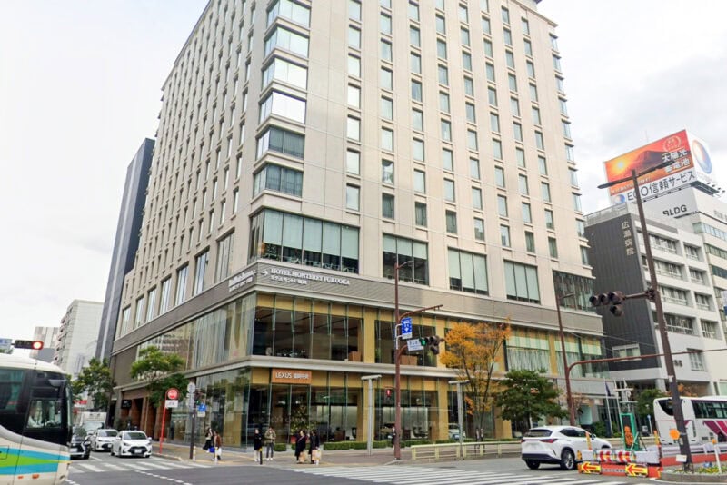 A modern multi-story building with large windows stands on a busy city intersection near Fukuoka hotels. Pedestrians cross the street, vehicles wait at the light, and shops, including LOUIS GARNEAU, occupy the ground floor. Japanese signs are visible.