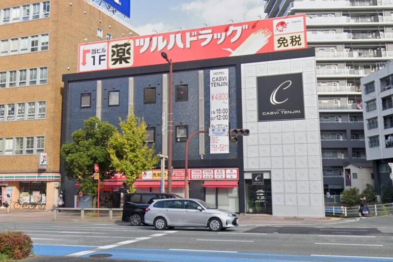 Street view of a city intersection with cars waiting at a traffic light, several mid-rise buildings, and storefronts including a drugstore and Hotel Casvi Tenjin—one of the well-known Fukuoka Hotels—with prominent signage in Japanese and English.