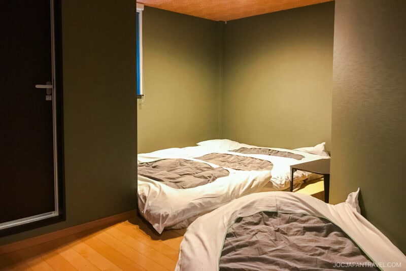 A cozy room at a Fukuoka Hotel, featuring three futons on a wooden floor with white bedding and gray blankets, set against green walls and a closed door.