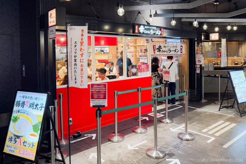A Japanese ramen restaurant, Ramen Runway Fukuoka - Kochan Ramen, features a striking red and black exterior, large windows, and people ordering at the counter. Menu signs and a queue with social distancing markers are visible in the indoor mall setting.