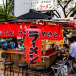 Fukuoka Yatai (屋台) – Street Food Stalls at the Heart of Fukuoka’s Nightlife A small open-air ramen stall, inspired by Fukuoka Yatai, with red lanterns and banners featuring Japanese text. Customers sit on stools at the counter, cooking utensils are visible, and the stall sits on a city sidewalk with trees and buildings nearby.