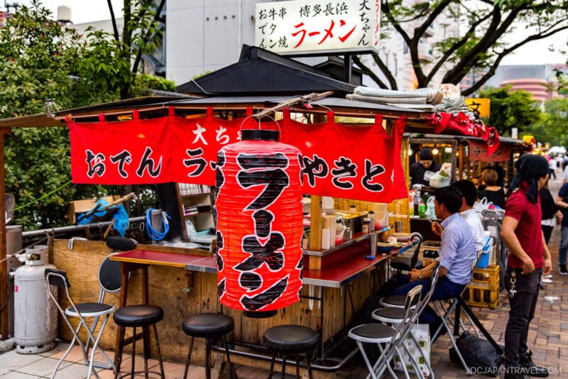 A small open-air ramen stall, inspired by Fukuoka Yatai, with red lanterns and banners featuring Japanese text. Customers sit on stools at the counter, cooking utensils are visible, and the stall sits on a city sidewalk with trees and buildings nearby.