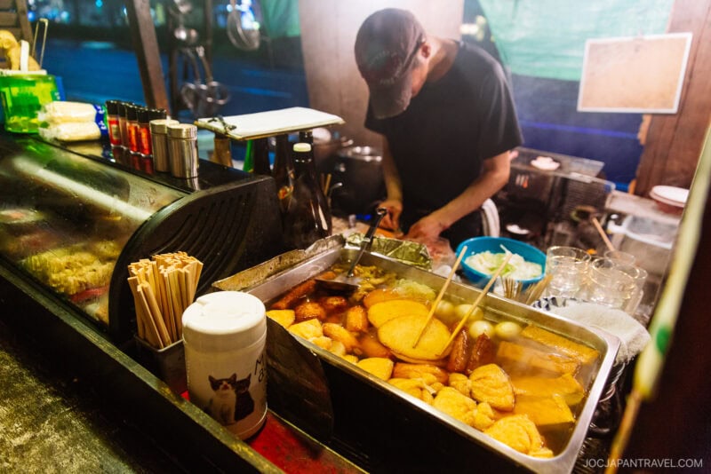 A street food vendor prepares food behind a Fukuoka Yatai stall, with steaming trays of assorted oden ingredients, chopsticks, condiments, and a container with a cat design visible in the foreground.