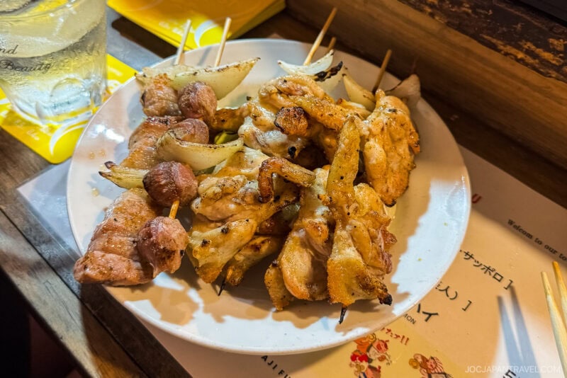 A plate of grilled skewers with chicken, onions, and meatballs is served on a table next to a glass of water and a yellow menu, capturing the lively street food vibes of Fukuoka Yatai.