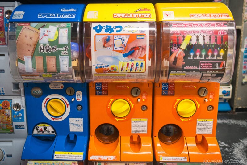 Three colorful Japanese capsule toy vending machines from Akihabara Gachapon Hall, each labeled in Japanese and displaying images of collectible toys like pouches, pens, and figurines.
