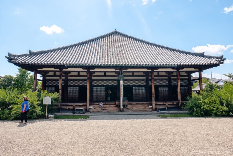 A traditional Japanese temple, Nara Gango-ji, with a tiled roof, wooden pillars, and open veranda, is surrounded by greenery. A person stands to the left, facing the building under a bright blue sky with scattered clouds.