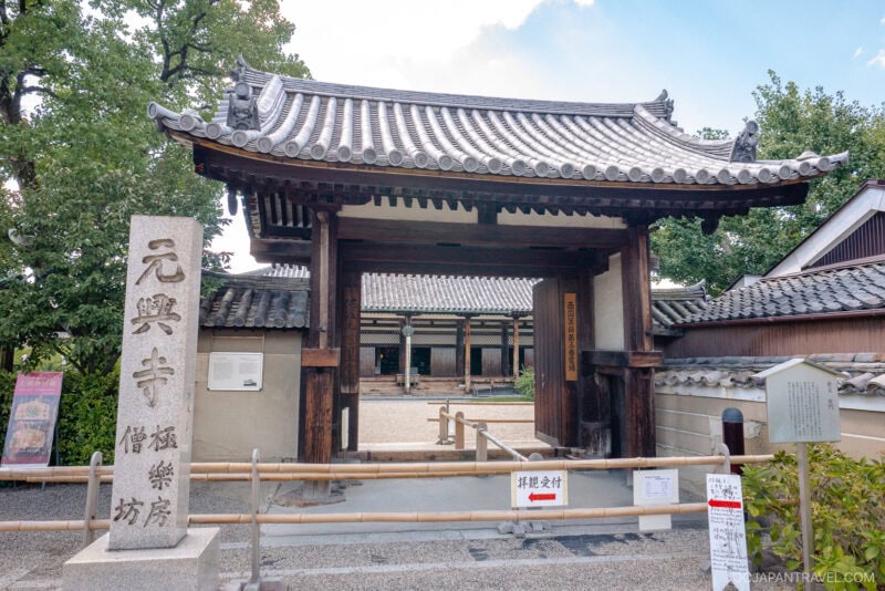Traditional wooden gate with tiled roof at Nara Gango-ji temple entrance, flanked by a stone marker with Japanese inscriptions. Pathway leads to a courtyard and additional temple buildings in the background.