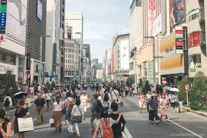 A busy city street in Japan, like Ginza Hokosha Tengoku, is filled with people walking, shopping, and enjoying the day. Tall buildings with colorful signs and billboards line both sides of the street under a partly cloudy sky.