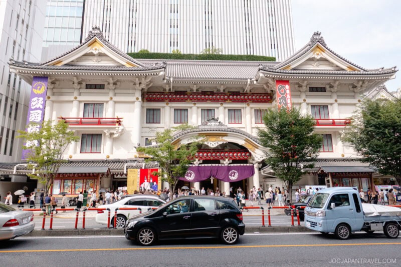 The Ginza Kabuki-za Theater showcases traditional Japanese architecture with ornate details, purple drapery, and decorative banners. People gather outside while modern cars line the street, set against a backdrop of towering buildings.