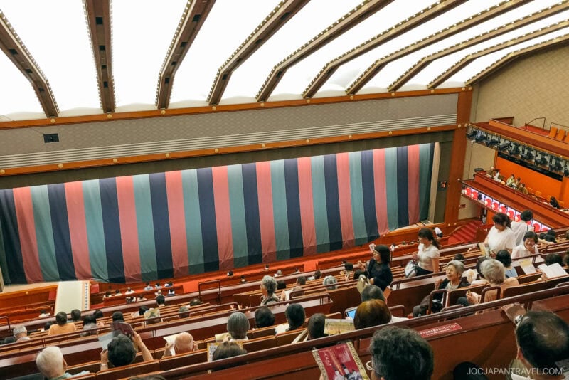 Audience members sit and wait inside the Ginza Kabuki-za theater with a striped curtain covering the stage. The theater has multiple seating levels and a modern ceiling. People are chatting and holding programs.
