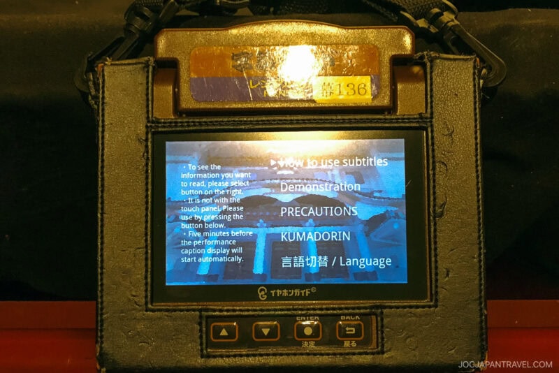 A handheld device at Ginza Kabuki-za displays instructions for using subtitles at the theater, with buttons for demonstration, precautions, language, and Kumadorin. A strap is attached to the device.