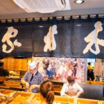 Ginza Kimuraya (木村屋總本店) – Home of Japan’s First Anpan A Japanese food stall with staff preparing dishes behind a counter, under a large navy curtain with bold white kanji characters. Customers stand in front, and a colorful mural decorates the back wall, evoking the spirit of Ginza Kimuraya.