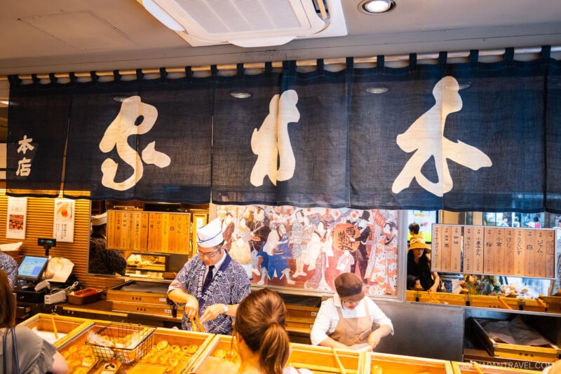 A Japanese food stall with staff preparing dishes behind a counter, under a large navy curtain with bold white kanji characters. Customers stand in front, and a colorful mural decorates the back wall, evoking the spirit of Ginza Kimuraya.