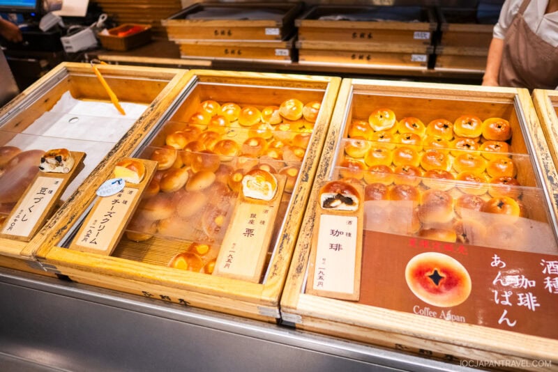 Display case with various types of Japanese bread buns from Ginza Kimuraya, each with different fillings, neatly arranged and labeled in Japanese. The golden brown buns are organized in wooden trays behind a glass cover.