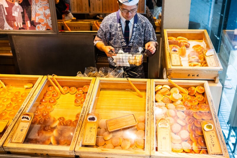 A shopkeeper in traditional Japanese attire arranges trays of assorted bread and pastries behind a glass counter in Ginza Kimuraya bakery. Wooden labels with Japanese writing identify each baked item.
