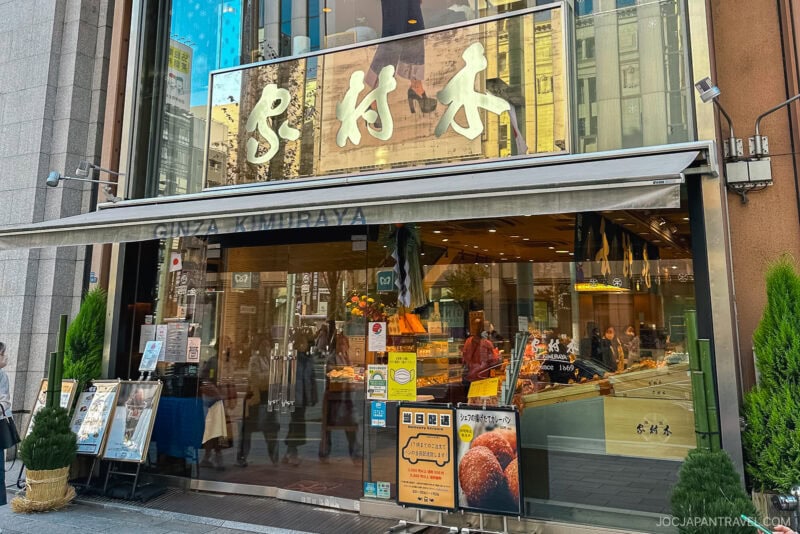 Street view of Ginza Kimuraya bakery in Tokyo with large glass windows, a Ginza Kimuraya sign with Japanese characters, greenery outside, and various breads visible inside the store.