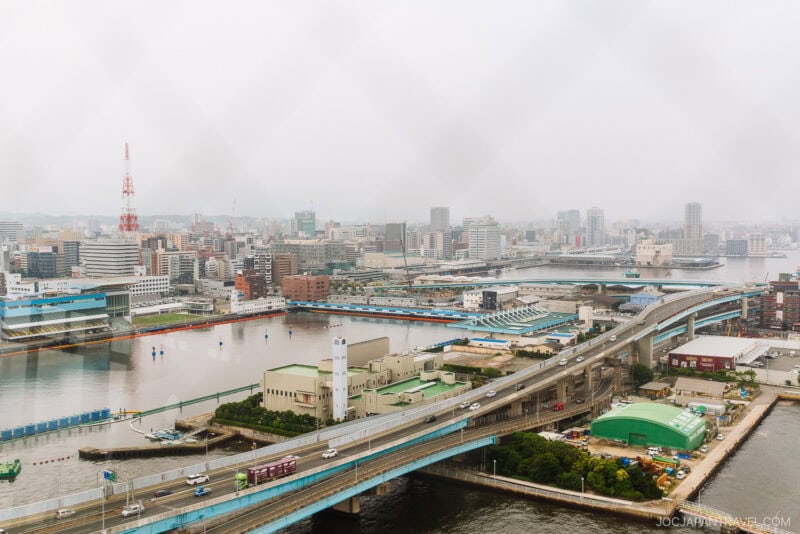 A cityscape view of Osaka, Japan, showing highways, bridges, and river channels on a cloudy day—seen through a window with a faint diamond-patterned grid—recalls the sweeping vistas from Hakata Port Tower.