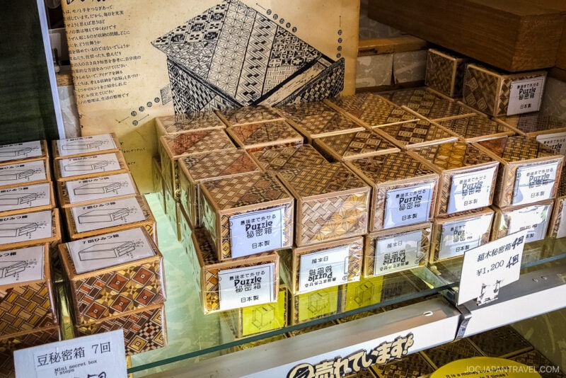 Wooden Hakone Himitsu-Bako Japanese puzzle boxes with intricate geometric patterns are displayed on a shelf. Some boxes have labels and price tags, while an illustrated instruction sheet is visible behind them.