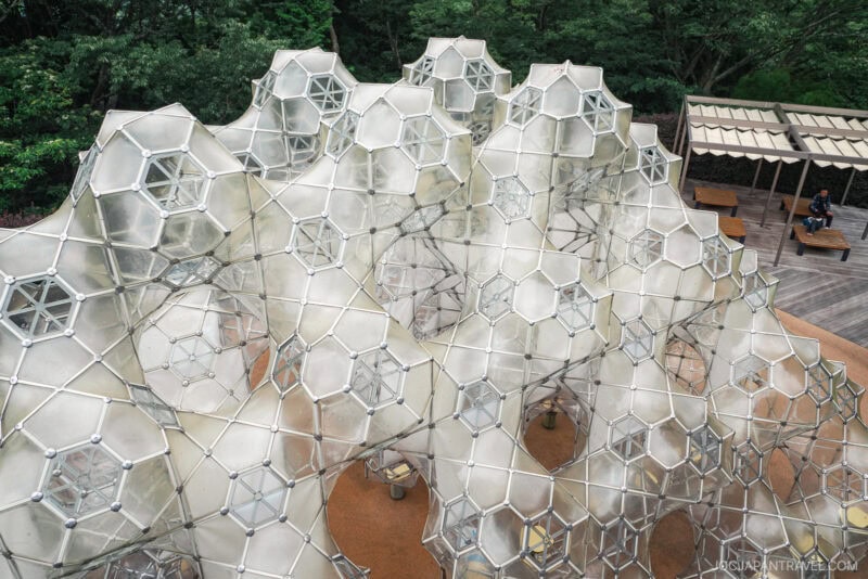A large geometric outdoor sculpture made of interconnected transparent hexagonal and pentagonal panels stands on a terrace surrounded by greenery at the Hakone Open-Air Museum, with benches and a person sitting nearby.