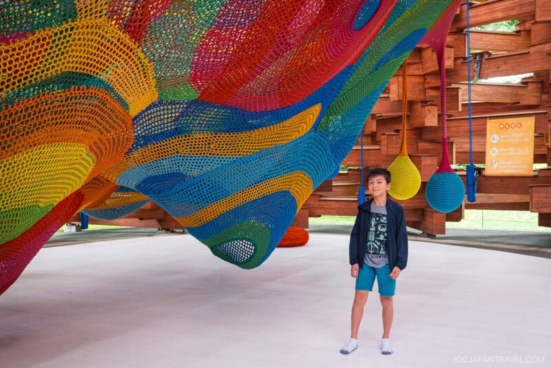 A boy in casual clothes stands under a large, colorful, netted playground structure indoors at the Hakone Open-Air Museum, surrounded by vibrant woven patterns and wooden walls.