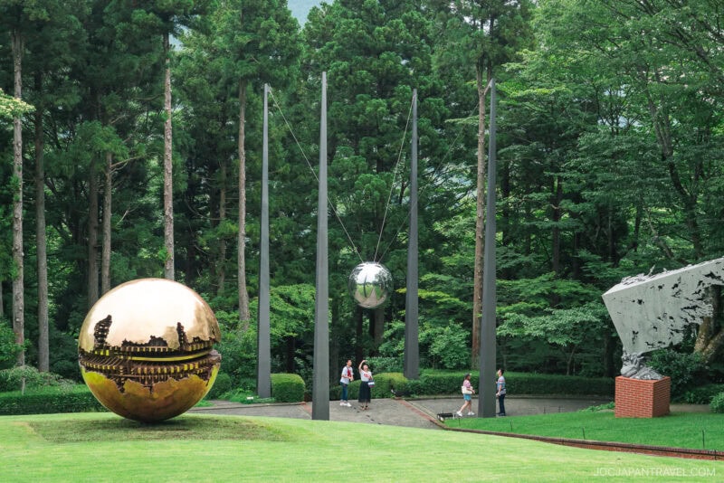 Sculptures of reflective silver and gold spheres stand amid tall trees and grass at the Hakone Open-Air Museum, while several people walk nearby in an outdoor park surrounded by greenery.