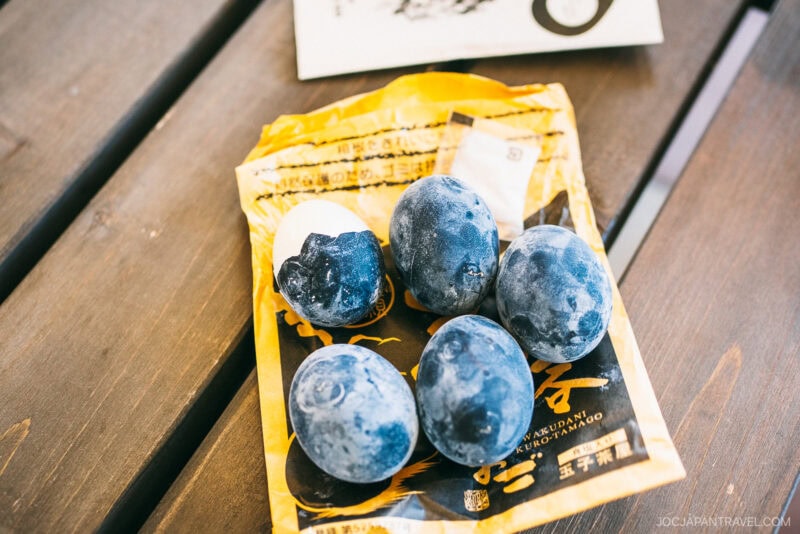 Six black eggs, some with cracked shells revealing white inside, rest on yellow packaging atop a wooden surface—delicacies from Hakone Ropeway’s famous Owakudani Valley.