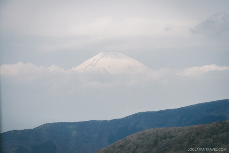Snow-capped Mount Fuji rises above a layer of clouds, with rolling hills and green forests in the foreground under a pale, cloudy sky near the Hakone Ropeway and Owakudani Valley.