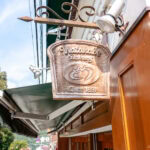Watanabe Bakery (渡邊ベーカリー): A Hidden Gem in Hakone’s Miyanoshita Village A wooden sign shaped like a bread slice hangs outside Hakone Watanabe Bakery, reading Watanabe Bakery Since 1897, with a loaf icon carved in the center. The brown and white building sits among trees in the background.