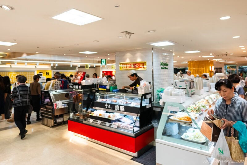 A busy food court in a brightly lit indoor market at Hankyu and Hanshin Department Stores Osaka, with people ordering and browsing various food stalls displaying prepared dishes behind glass counters.