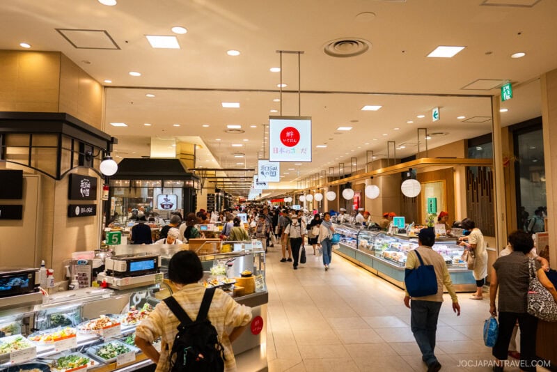 A busy Japanese supermarket inside Hankyu and Hanshin Department Stores Osaka, with shoppers walking through wide aisles lined with bright food displays, overhead signs, and modern lighting. People browse fresh and prepared foods in a clean, spacious setting.