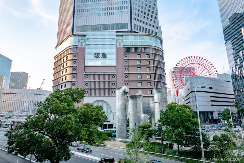 A large modern building with the Hankyu Department Store sign in Japanese, part of the Hankyu and Hanshin Department Stores Osaka, surrounded by cityscape, trees, busy streets, and a red Ferris wheel under a clear blue sky.
