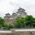 Himeji Castle (姫路城) – Japan’s White Heron Castle Himeji Castle is a white traditional Japanese castle with multiple roofs, standing on a stone foundation, surrounded by green trees and a stone wall under a cloudy sky.