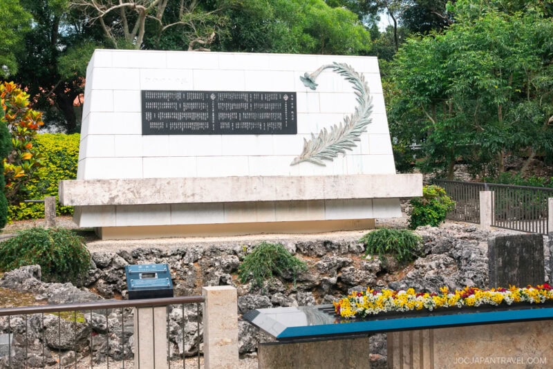 A white stone memorial monument with a black plaque listing names, accented by a metal olive branch design, stands near the Himeyuri Peace Museum. The monument is surrounded by greenery, flowers, and a small fence.