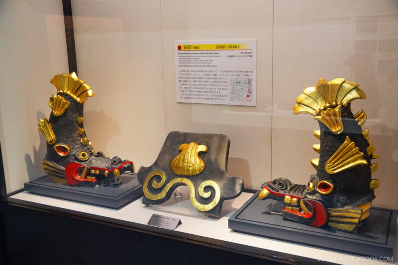 Two ornate, black and gold fish-shaped roof ornaments from Hiroshima Castle with red accents are displayed on either side of a gray, gold-trimmed roof tile in a museum case, with an informational sign in the background.