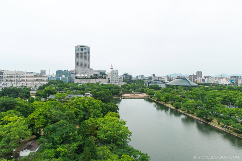 A cityscape view featuring a large park with dense green trees and a pond in the foreground, surrounded by modern buildings and Hiroshima Castle’s tall tower in the background under a cloudy sky.