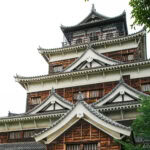 Exploring Hiroshima Castle 広島城 A close-up view of Hiroshima Castle showcases its multi-tiered wooden and white exterior, ornate rooflines, and intricate architectural details, with green trees partially framing this iconic Hiroshima Castle landmark.