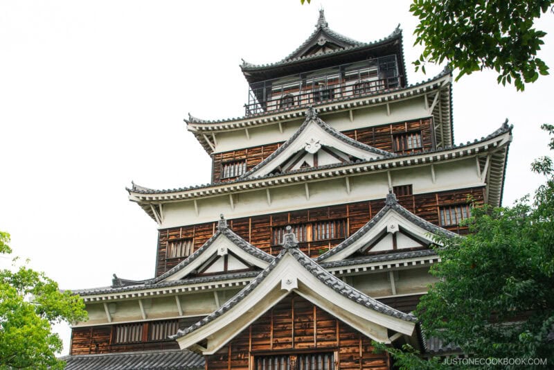 A close-up view of Hiroshima Castle showcases its multi-tiered wooden and white exterior, ornate rooflines, and intricate architectural details, with green trees partially framing this iconic Hiroshima Castle landmark.