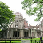 The Hiroshima Peace Memorial, a partially destroyed concrete building with a domed framework, stands surrounded by green trees and grass within Hiroshima Peace Memorial Park under a bright, cloudy sky.